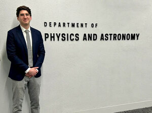 Scott Carmichael wearing a blue suit, grey pants standing next the Notre Dame Department of Physics and Astronomy sign.