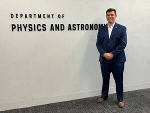 Thomas Bailey in a blue suit standing next the sign says Department of Physics and Astronomy