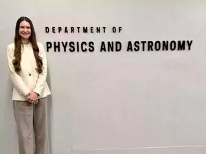 Mateny in white blazer jacket and khaki pants standing in front of the sign that says department of physics and astronomy