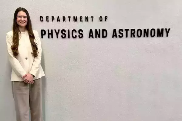 Miriam Mateny in white blazer jacket and khaki pants stands in front of the sign that says department of physics and astronomy