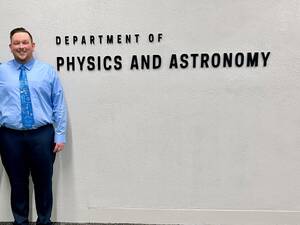 John McDonaugh wearing light blue dress shirt with a tie and dark navy pants stands in front of the sign that says department of physics and astronomy